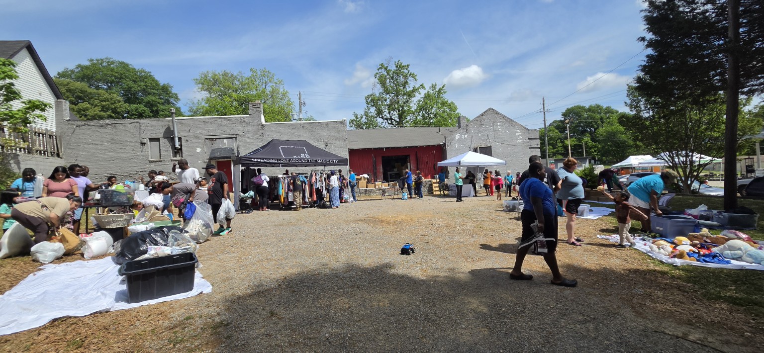 Families and volunteers gathering around clothing and essential-goods stations at a Be Kind Birmingham community day